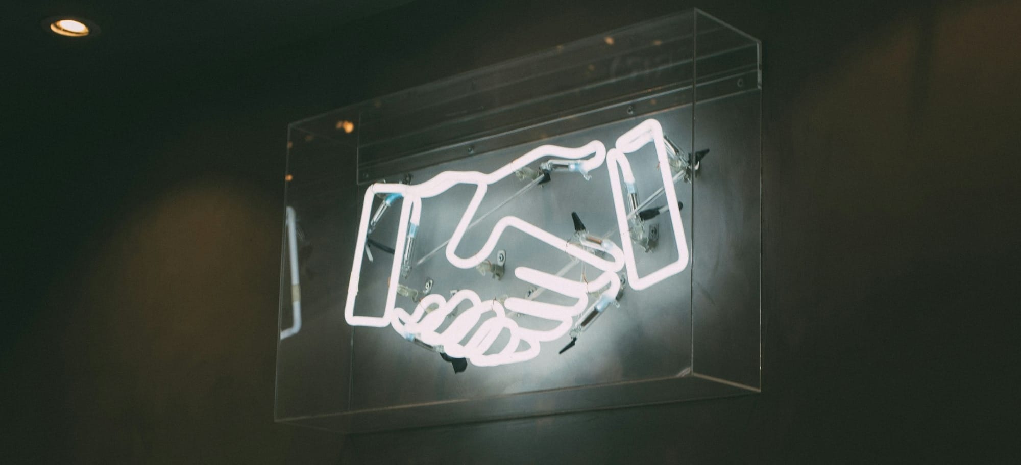 A neon sign of two shaking hands, behind a glass or plastic pane and with a moody, smoky lighting